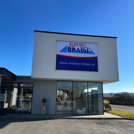 Ein weißes Gebäude mit einem Schild, das Elektro Braun und eine Website-Adresse anzeigt. Der Eingang hat Glastüren, die Waschmaschinen im Inneren zeigen.