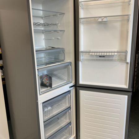 Ein offener Kühlschrank mit Glastüren und Regalen. Der Markenname Miele ist sichtbar. Das Innere ist leer mit sichtbaren Schubladen und Fächern.