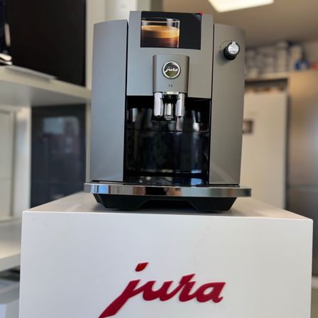 Eine Jura E6 Espressomaschine wird auf einem weißen Podest mit dem Jura-Logo präsentiert. Die Maschine hat eine Glastasse in ihrem Halter.
