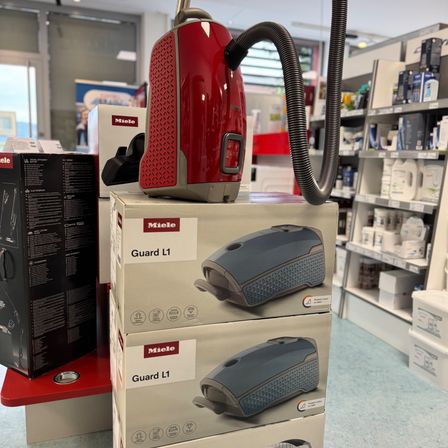 Mehrere Kartons eines roten und grauen Miele-Staubsaugers sind in einem Geschäft ausgestellt. Dahinter befinden sich Regale mit verschiedenen Reinigungsprodukten.