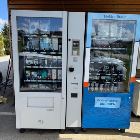 Zwei weiße und blaue Verkaufsautomaten mit Glastüren stehen draußen. Der linke Automat hat die Aufschrift PUSH und verschiedene Artikel im Inneren. Der rechte Automat hat die Aufkleber Elektro Braun und Produkt-Ausgabe Drucken.