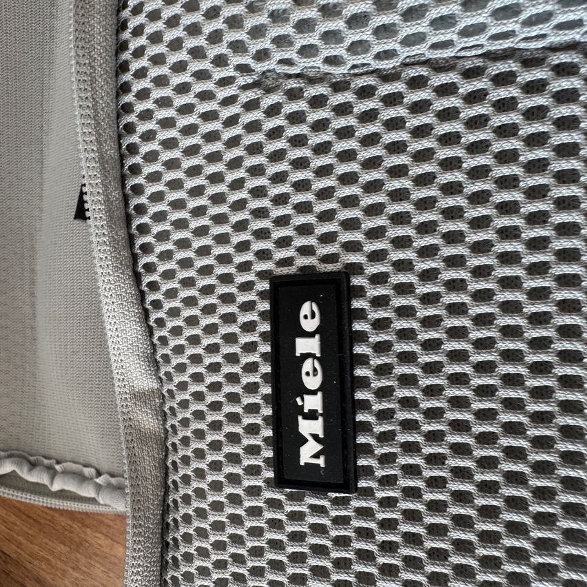 Nahaufnahme eines grauen Netzgewebes mit einem schwarzen Miele-Etikett in der Mitte. Das Gewebe hat ein kleines Lochmuster. Das Bild ist aus einer leicht schrägen Perspektive aufgenommen.