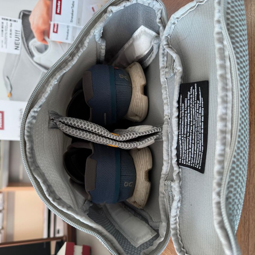 Eine offene Reisetasche mit einem Fach für Schuhe. Im Inneren befinden sich zwei Paar Sneaker. Die Tasche hat Fächer und ein Etikett mit Text und Zahlen.