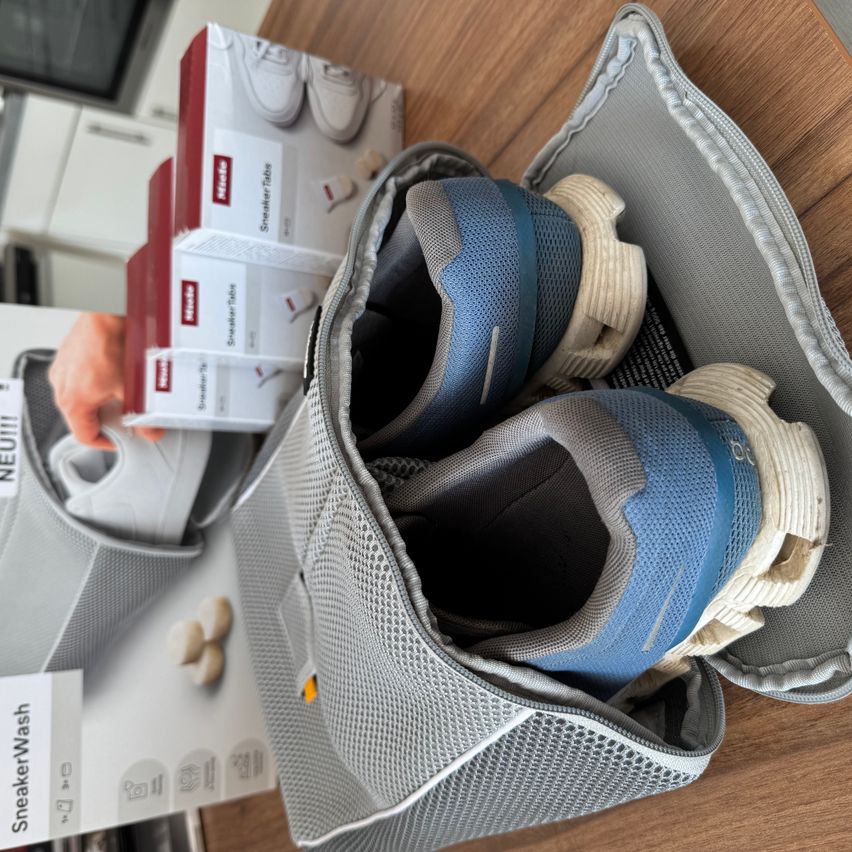 Ein Paar blaue und graue Turnschuhe befindet sich in einer Netztasche. Neben der Tasche sind zwei Schachteln eines Produkts namens SneakerTube.