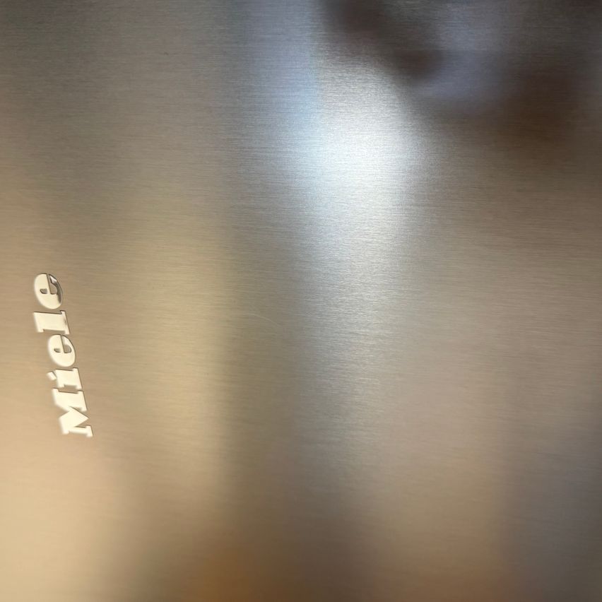 Ein Nahaufnahme einer Edelstahloberfläche zeigt das Wort 'Miele' deutlich. Die Oberfläche reflektiert Licht und deutet auf eine polierte Oberfläche hin.