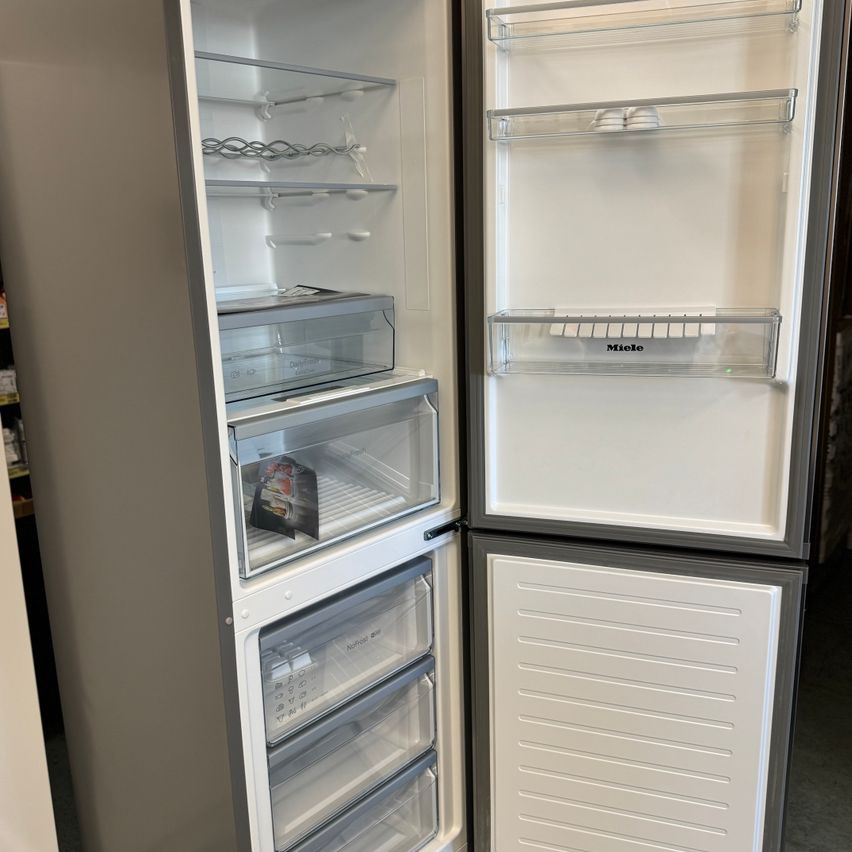 Ein leerer Miele-Kühlschrank mit offenen Türen, der transparente Schubladen, Regale und Fächer zeigt. Der Gefrierschrank befindet sich links und der Kühlschrank rechts.