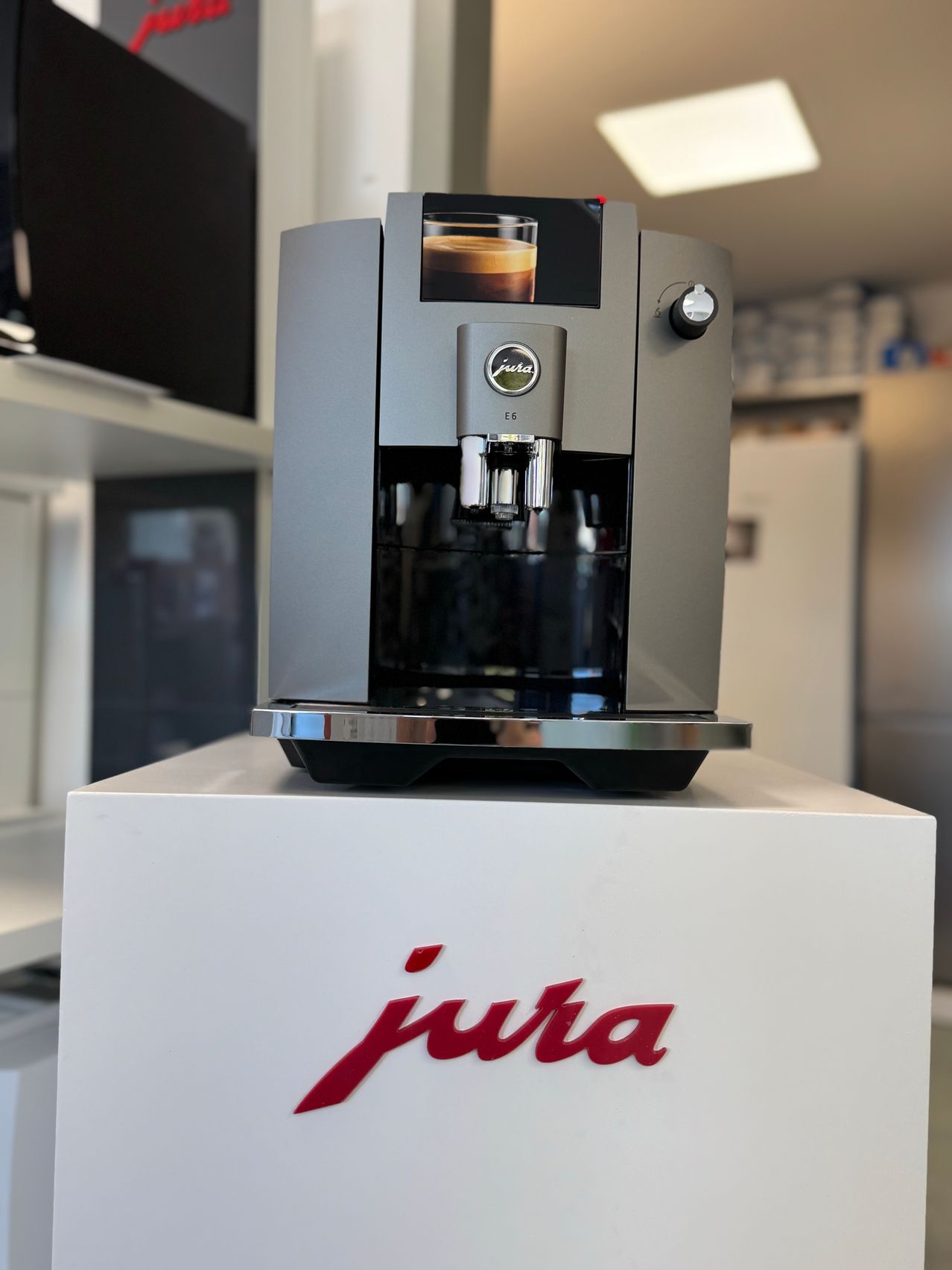 Eine Jura-Kaffeemaschine Modell E6 ist auf einem weißen Podest ausgestellt, wobei das Jura-Logo deutlich auf dem Podest zu sehen ist.