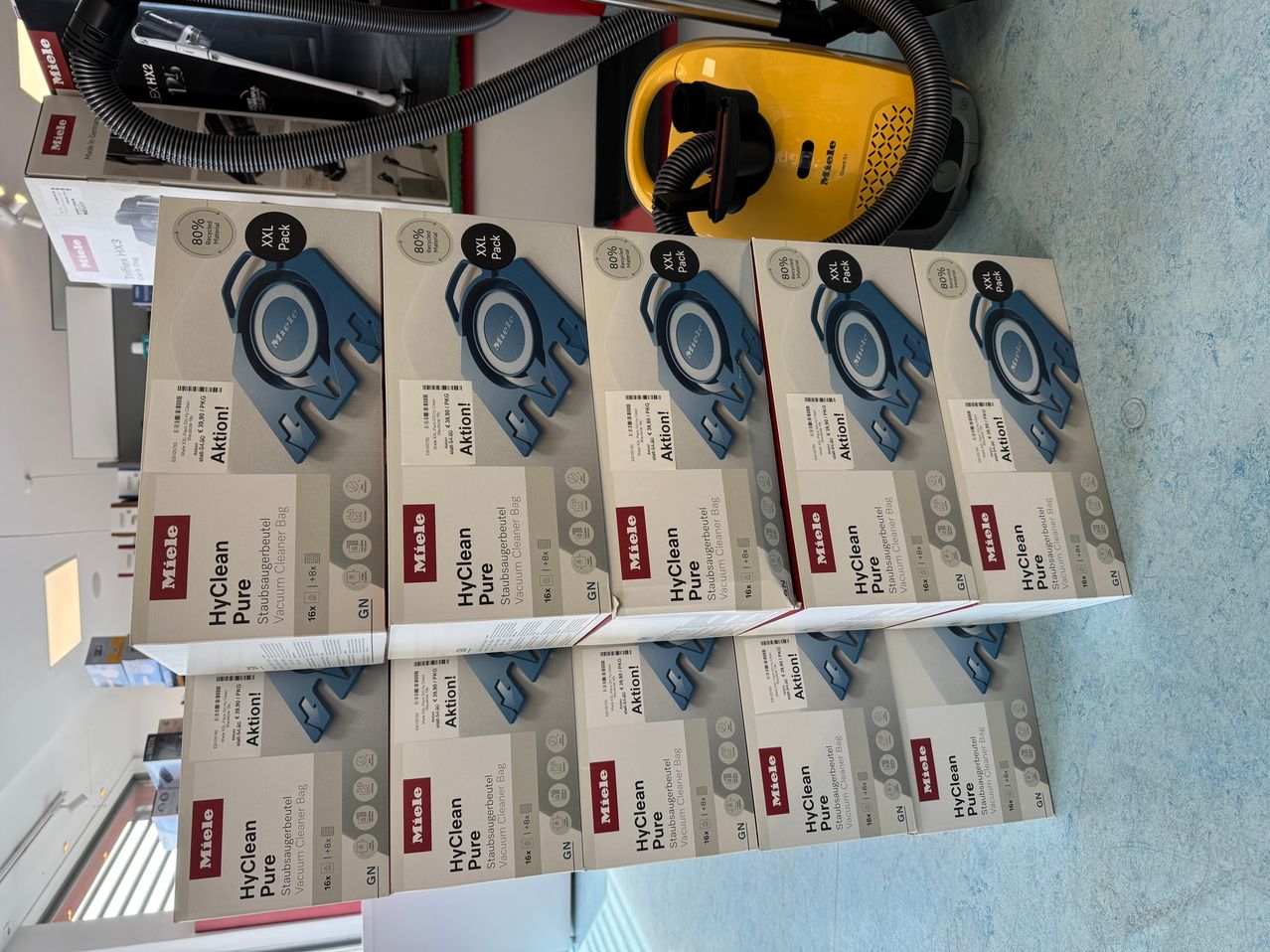 Stack of Miele HyClean Pure vacuum cleaner bags. The bags are labeled with the brand name and the product details. There is a yellow vacuum cleaner in the background.