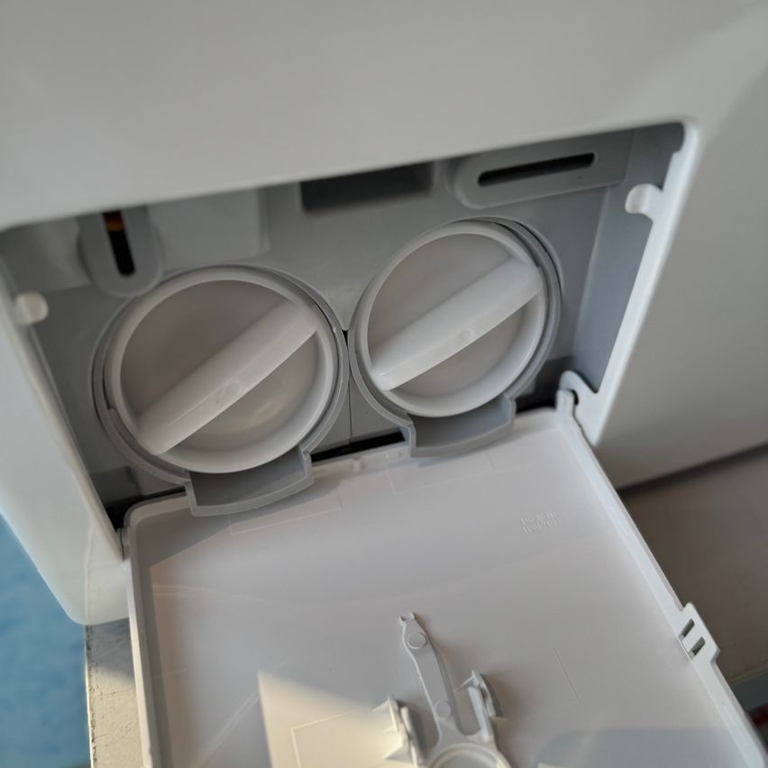 Eine Nahaufnahme eines geöffneten weißen Geräts, wahrscheinlich ein Geschirrspüler, mit geöffneter Tür und Fächern, die die internen Komponenten und ein kleines weißes Objekt im Inneren zeigen.