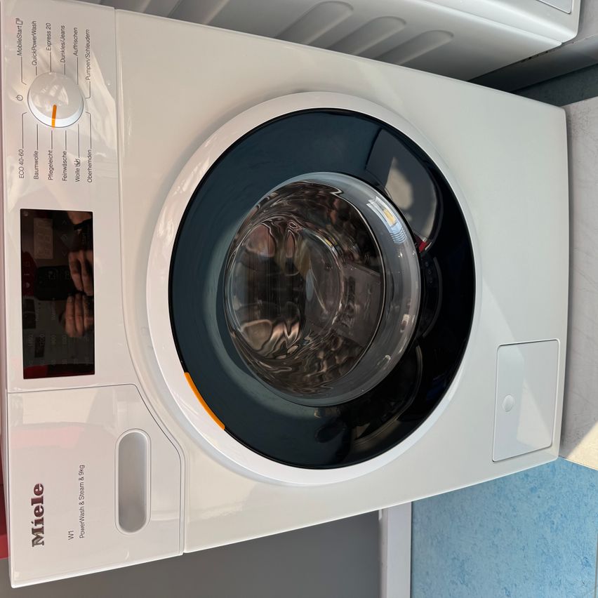 Eine weiße Frontlader-Waschmaschine mit digitalem Display ist zu sehen. Die Maschine hat mehrere Knöpfe und Drehregler für verschiedene Waschfunktionen.