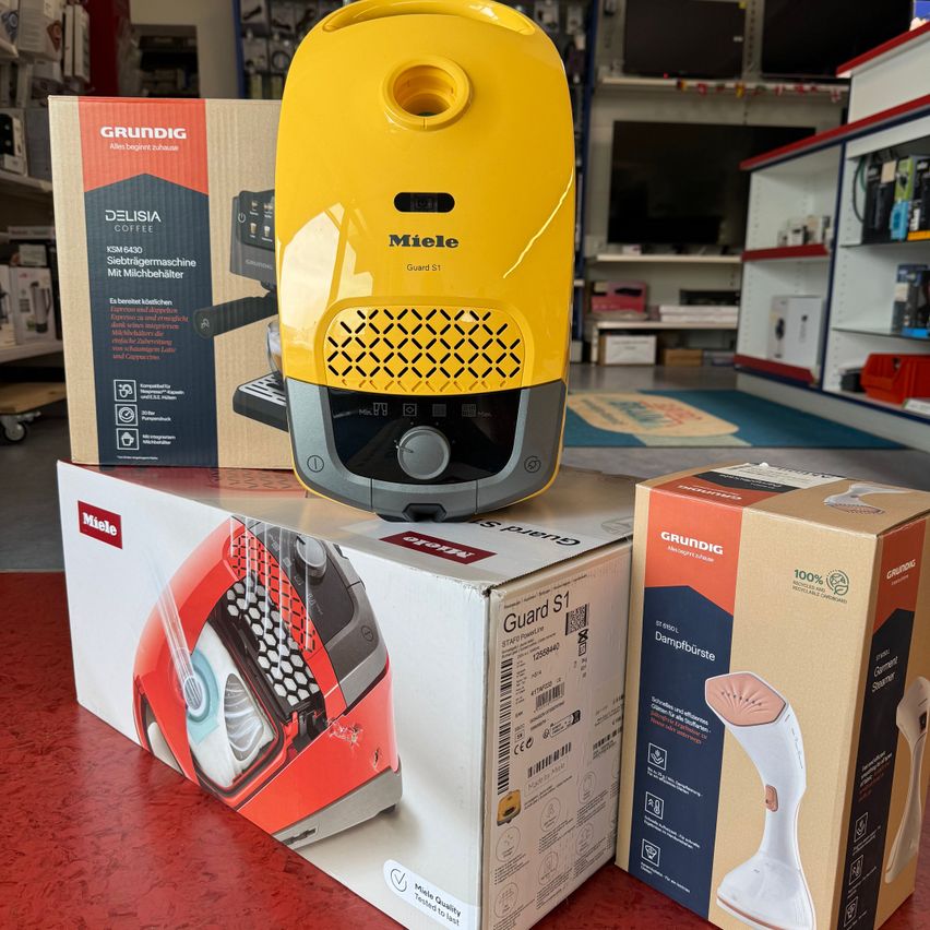 Ein gelber und grauer Miele-Staubsauger wird auf einem weißen Kasten mit einem roten und grauen Bild des Staubsaugers ausgestellt. Daneben befindet sich eine Box mit Grundig-Kaffee.