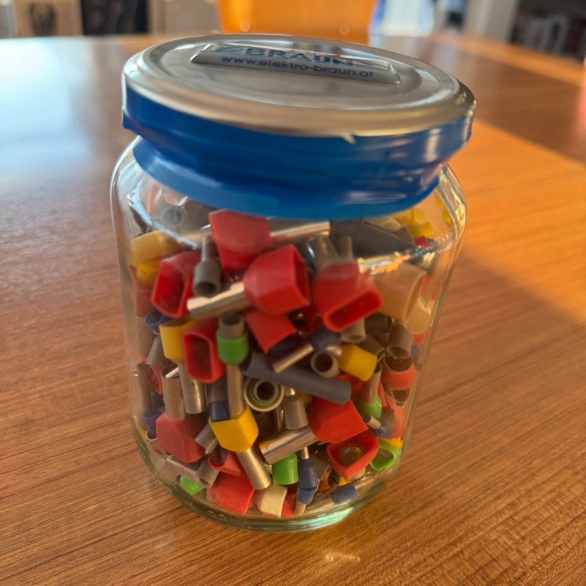 Ein Glasglas mit verschiedenen farbigen Kunststoffverbindern und einem blauen Deckel mit einer Aufschrift, die 'ZEBRAUN www.elektro-braun.at' auf einem Holztisch liest.