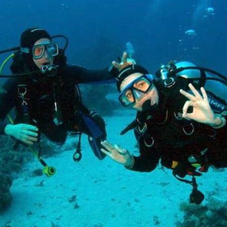 Zwei Taucher unter Wasser, einer trägt einen schwarzen Tauchanzug und der andere einen schwarzen Tauchanzug mit einer weißen Uhr, beide machen Friedenszeichen. Sie sind von Korallenriffen umgeben.