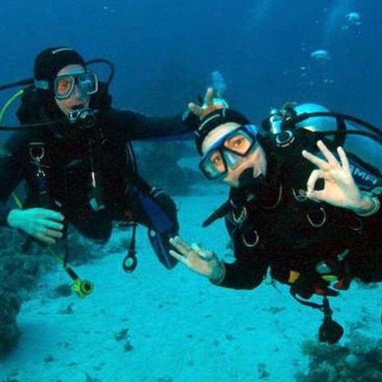 Zwei Taucher unter Wasser, einer trägt einen schwarzen Tauchanzug und der andere einen schwarzen Tauchanzug mit einer weißen Uhr, beide machen Friedenszeichen. Sie sind von Korallenriffen umgeben.
