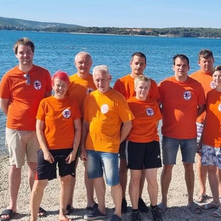 Eine Gruppe von Menschen in orangefarbenen Shirts posiert für ein Foto an einem Kiesstrand mit einer Wasserfläche und einem hügeligen Hintergrund.
