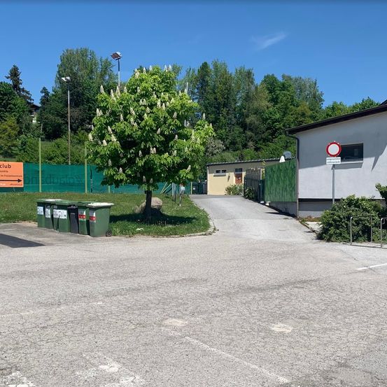 Ein leerer Parkplatz mit grünen Mülleimern, einem Baum und einem Schild ist von einem Zaun umgeben. Ein Gebäude mit einem Verkehrsschild befindet sich rechts.