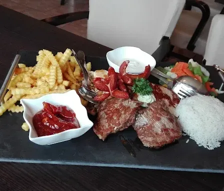 Ein schwarzer Schieferteller mit Steak, Reis, Pommes, Gemüse, Saucen und Besteck auf einem Holztisch.