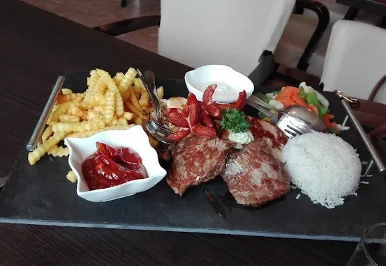 Ein schwarzer Schieferteller mit Steak, Reis, Pommes, Gemüse, Saucen und Besteck auf einem Holztisch.
