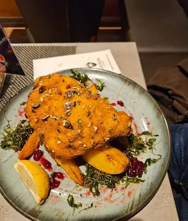 Ein Teller mit paniertem Fisch, Karotten, grünen Kräutern und einer Zitronenscheibe, mit einem Menü und einer Serviette im Hintergrund.