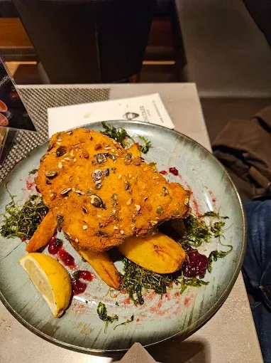 Ein Teller mit paniertem Fisch, Karotten, grünen Kräutern und einer Zitronenscheibe, mit einem Menü und einer Serviette im Hintergrund.