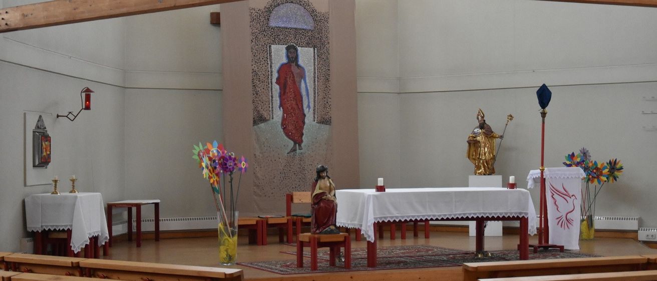 Innenansicht einer Kapelle mit einem großen Gemälde von Jesus an der Wand. Eine kleine Statue einer Frau sitzt auf einer Holzplattform. Ein Tisch mit Kerzen steht vor dem Gemälde. Eine Statue eines Heiligen mit einem Stab befindet sich auf der rechten Seite.