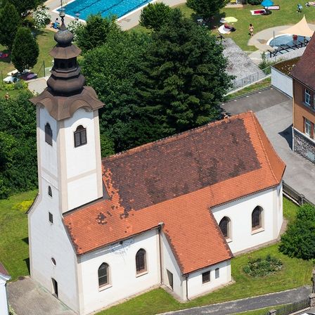 Luftaufnahme einer Kirche mit braunem Dach und hohem Turm, umgeben von einem grünen Rasen und Bäumen. In der Nähe befindet sich ein Schwimmbad mit schwimmenden Personen.
