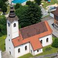 Luftaufnahme einer Kirche mit braunem Dach und hohem Turm, umgeben von einem grünen Rasen und Bäumen. In der Nähe befindet sich ein Schwimmbad mit schwimmenden Personen.