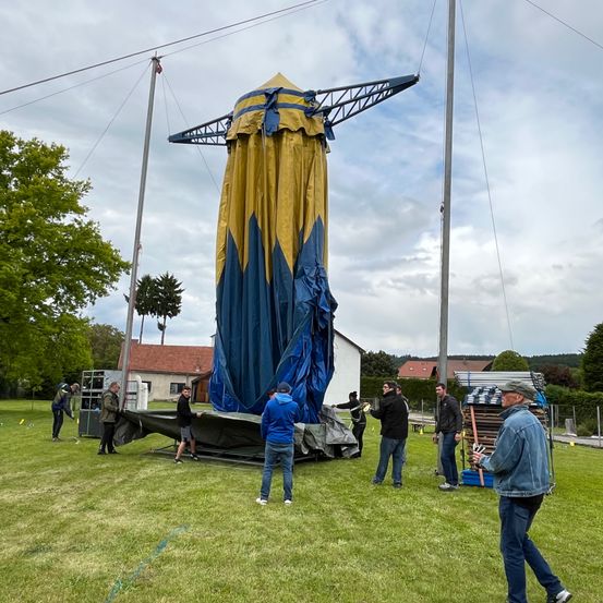 Mehrere Personen bauen ein großes blau-gelbes Zelt auf einem Rasenfeld mit bewölktem Himmel auf. Bäume und Gebäude sind im Hintergrund.