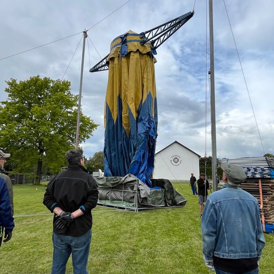 Ein Kran hebt ein großes blaues und gelbes Zelt auf einem Rasenfeld. Mehrere Leute beobachten, darunter einer in einer Jeansjacke. Ein Haus und ein Stapel Holz sind im Hintergrund.
