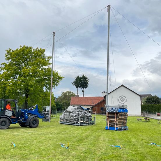 Ein blauer Traktor fährt auf einem Feld mit zwei Masten. Dahinter befinden sich ein Holzhaufen und eine schwarze Plane. Ein Haus mit rotem Dach ist im Hintergrund.