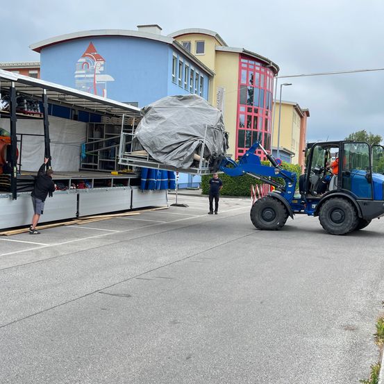 Ein großer Gegenstand wird von einem blauen Traktor in einem städtischen Umfeld angehoben, mit Arbeitern in der Nähe und einem Gebäude mit einem Wandbild im Hintergrund.