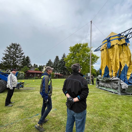Drei Männer in Jacken stehen auf einem Rasen mit einem großen gelben und blauen Zelt, das teilweise aufgebaut ist. Hinter ihnen befindet sich ein Maschinenfahrzeug und ein Haus.
