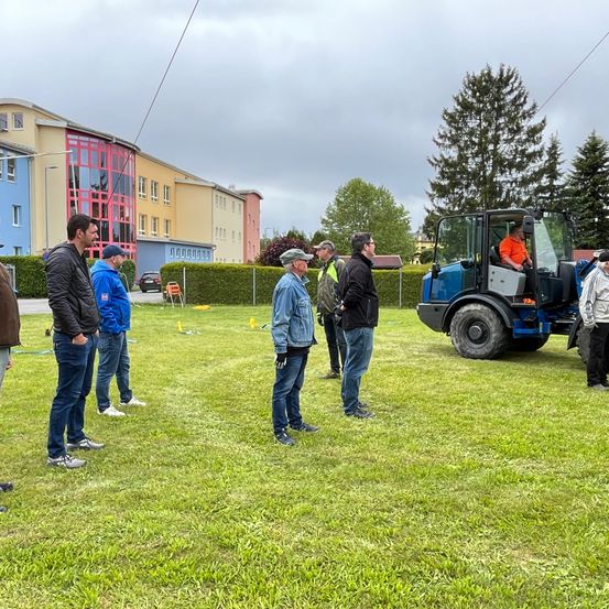 Eine Gruppe von Männern in Jacken und Mützen steht auf einem Rasen und diskutiert etwas. Hinter ihnen ist ein blauer Traktor geparkt. Im Hintergrund befinden sich Gebäude, Bäume und ein geparktes Auto.