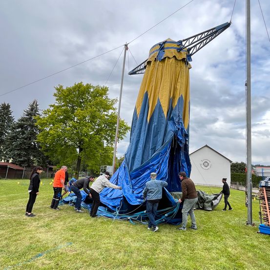 Eine Gruppe von Menschen arbeitet zusammen, um ein großes Zelt auf einem Feld aufzubauen. Das Zelt ist blau und gelb.
