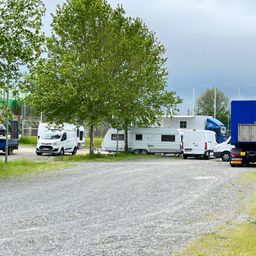 Ein paar Lieferwagen und Wohnwagen stehen auf einem Schotterplatz in der Nähe eines Baumes, mit einem blauen Lastwagen im Hintergrund.
