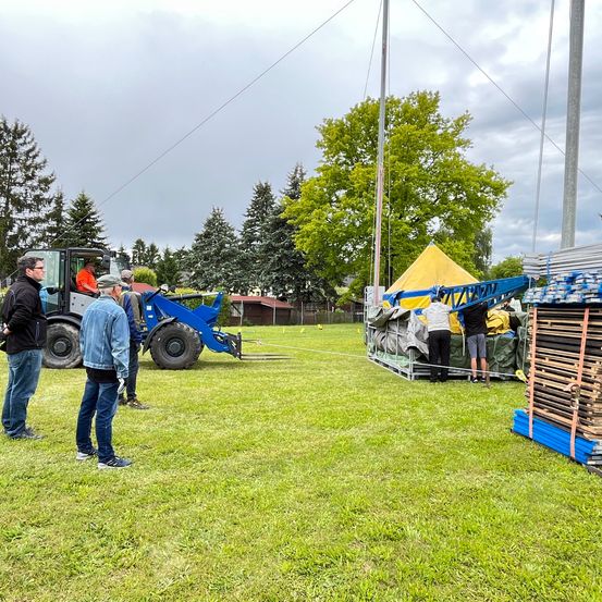 Eine Gruppe von Menschen steht auf einem Feld mit einem Traktor und einem gelben Zelt. Ein Mann fährt den Traktor, während andere eine blaue Kran entladen. Das Gebiet ist von Bäumen und Gebäuden umgeben.