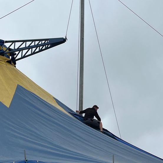 Ein Mann arbeitet unter einem bewölkten Himmel an einem blauen und gelben Zelt, sitzt am Rande des Zeltes und hält eine Seil fest.