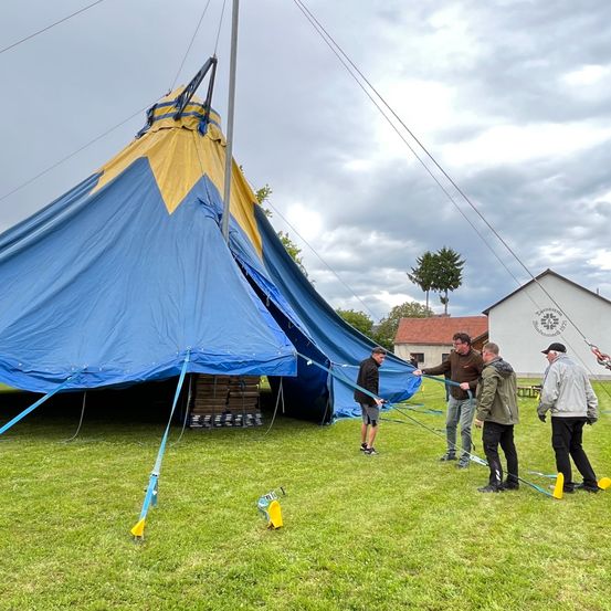 Eine Gruppe von Menschen richtet ein großes blau-gelbes Zelt auf einem Rasenfeld unter einem bewölkten Himmel auf.