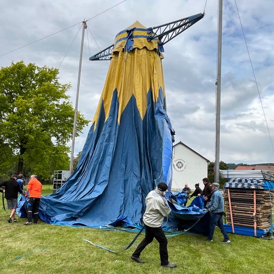 Eine Gruppe von Menschen baut ein großes blaues und gelbes Zelt auf einer Wiese auf. In der Nähe sind Holzpaletten und ein Haus zu sehen.