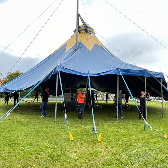 Ein großes blaues und gelbes Zelt wird auf einer Rasenfläche aufgebaut. Mehrere Personen arbeiten am Zelt und sichern es mit Seilen und Stangen. Der Himmel ist bewölkt.