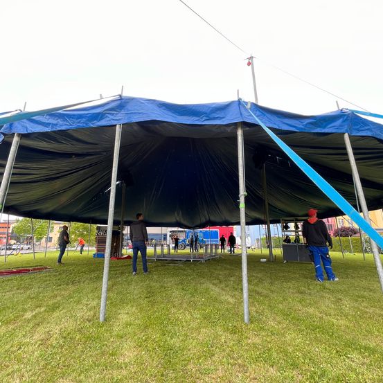 Ein blaues Zelt wird auf einem Rasenfeld aufgebaut, wobei mehrere Personen daran arbeiten. Einige Personen stehen in der Nähe des Zeltes, während andere an der Ausrüstung arbeiten. Im Hintergrund sind Gebäude und Autos zu sehen.