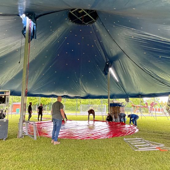 Eine Gruppe von Menschen arbeitet zusammen, um ein Zelt auf einem Rasenfeld aufzubauen. Ein Mann steht und beobachtet, während andere knien und den roten Boden im Inneren anpassen.