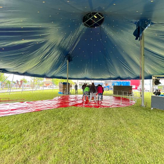 Unter einem großen blauen Zelt stehen mehrere Personen in einem Kreis auf einer roten Matte. Das Zelt hat eine kreisförmige Öffnung oben. Das Gelände ist von einer Metallbarriere umgeben.
