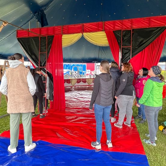 Eine Gruppe von Menschen steht in einem Zirkuszelt mit roten und schwarzen Vorhängen. Einige schauen zur Decke, andere zum Eingang. Eine Leiter ist links sichtbar.