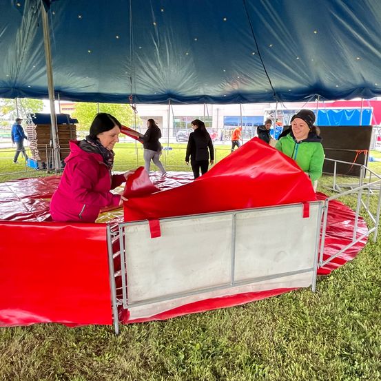 Zwei Frauen bauen eine rote Struktur unter einem blauen Zelt auf. Eine Frau hält die rote Abdeckung, während die andere das Metallgitter justiert. Dahinter gehen einige Leute auf einem Rasenfeld.