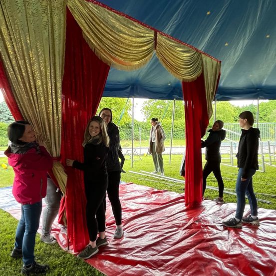 Fünf Frauen richten ein Zelt mit roten und goldenen Vorhängen auf. Das Zelt ist blau mit Sternen. Eine Frau macht ein Foto auf der rechten Seite. Bäume und ein Zaun sind im Hintergrund.