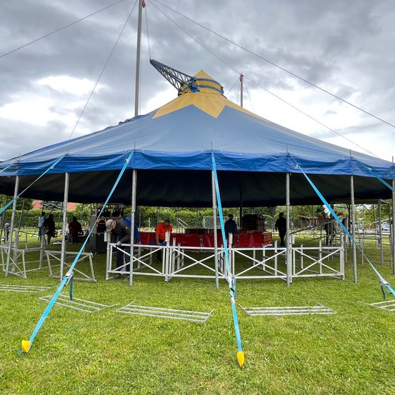 Ein großes blaues und gelbes Zirkuszelt wird auf einem Rasenfeld aufgebaut. Mehrere Personen arbeiten am Zelt, und einige stehen in der Nähe. Das Zelt ist von Metallrahmen umgeben.