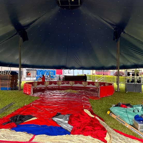 In einem großen Zelt steht eine rote ringförmige Struktur auf dem Rasen. Eine Person steht neben einem Fahrzeug. Einige Leute stehen hinter dem Zelt. Ein grüner Beutel und eine Metallbox sind neben dem Zelt platziert.