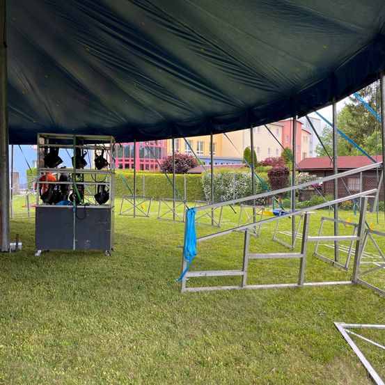 Ein großes Zelt auf einem Rasenfeld mit einer Metallstruktur und Ausrüstung darunter. Gebäude im Hintergrund.
