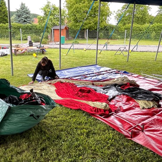 Eine Person kniet auf dem Rasen und ordnet Teile eines Heißluftballons auf dem Boden an. In der Nähe sind verschiedene Planen und Ausrüstungsgegenstände ausgebreitet. Bäume und ein Holzschuppen sind im Hintergrund zu sehen.
